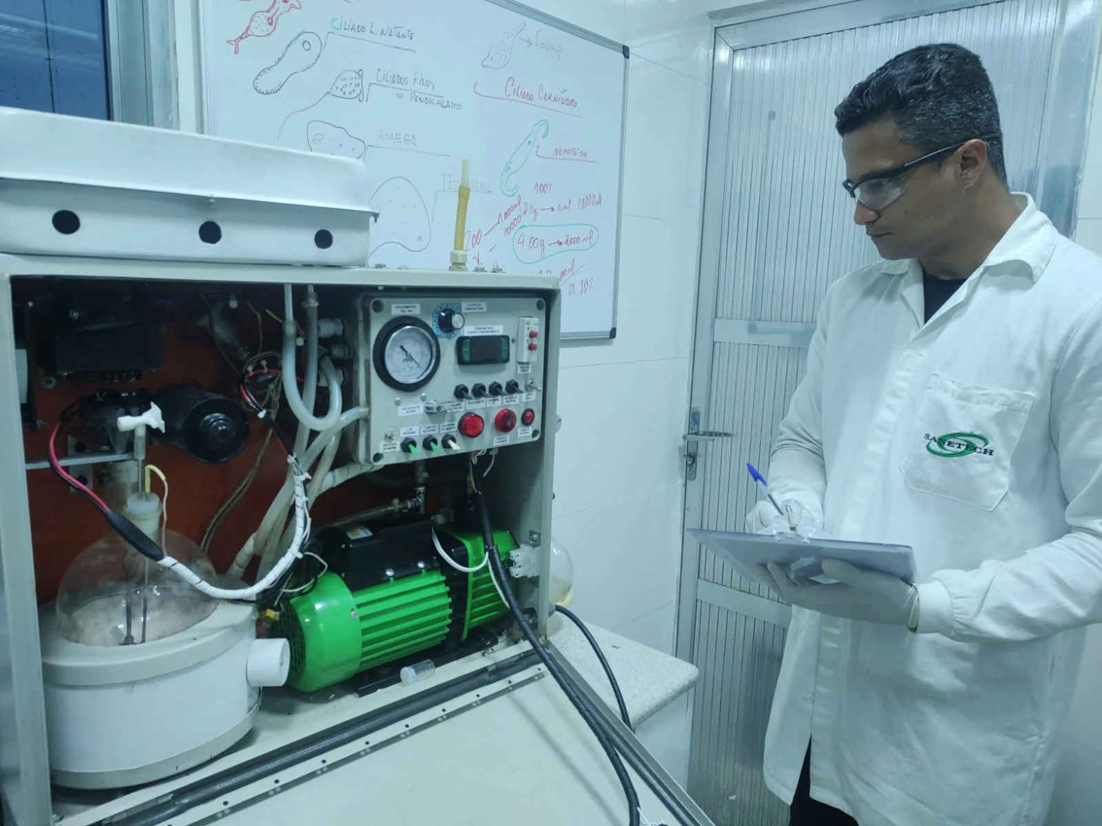 Técnico Sanetech examinando rota-evaporador em bancada de tratabilidade com diagrama de células microbiológicas no quadro