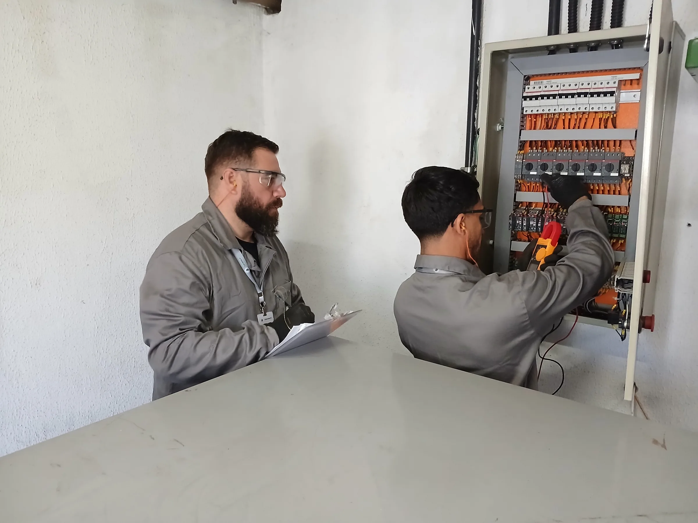 Dois técnicos: um anotando em clipboard, outro fazendo medição com multímetro em painel elétrico